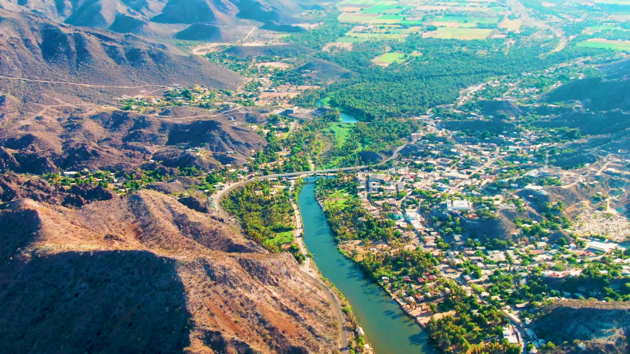Wide aerial view of Mulege with the river and town centered between dark hill shadows, green vegetation, distant fields, and no visible people.