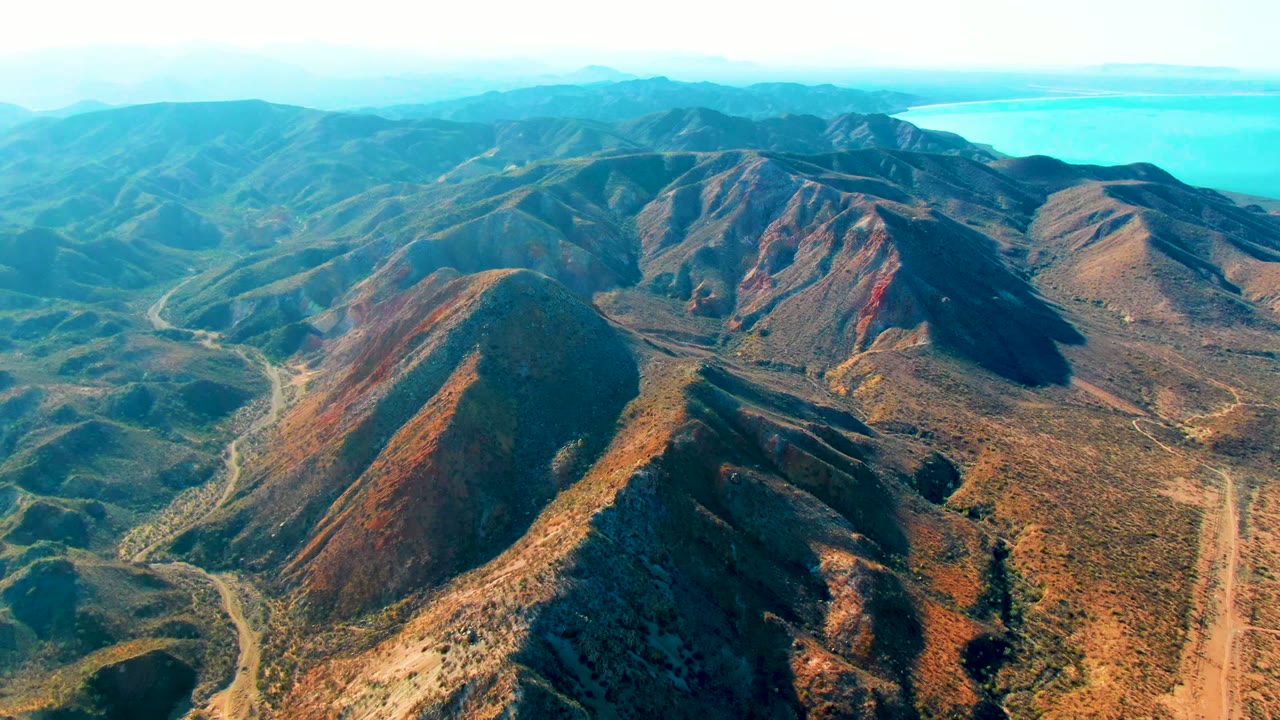 Opening aerial frame over rugged ridges near Mulege, Baja California Sur, with dry hills, valley tracks, distant blue-green water, and no visible people.