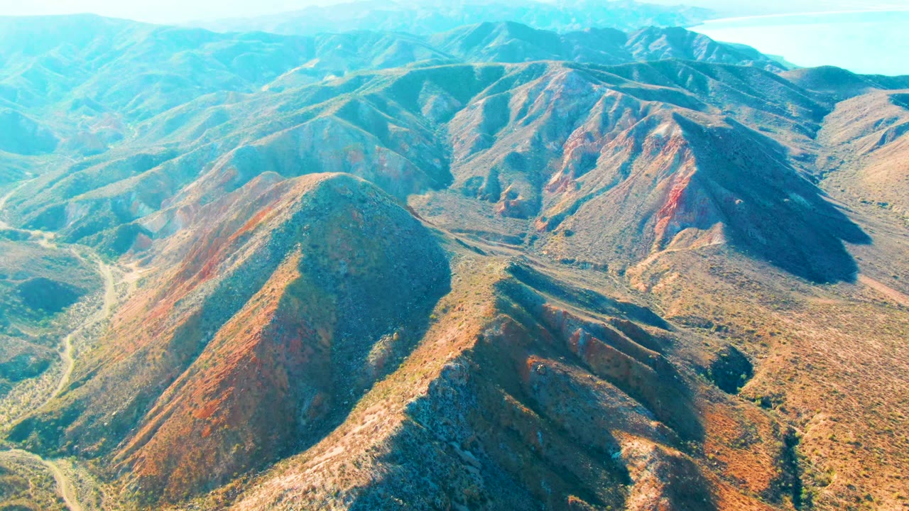 Aerial view over Mulege, Baja California Sur mountain ridges with a rounded foreground slope, pale road lines in the valley, distant hills, and no visible people.