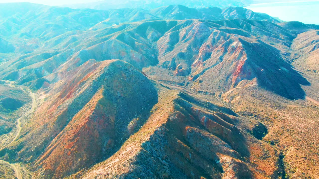 Aerial view of Mulege, Baja California Sur ridges with the foreground hill shifted slightly across the frame, road curves at left and center, hazy distant mountains, and no visible people.