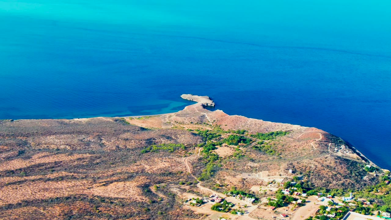 Opening aerial frame over Mulege, Baja California Sur, with deep blue ocean behind a dry rocky peninsula, scattered roads, greenery, and no visible people.