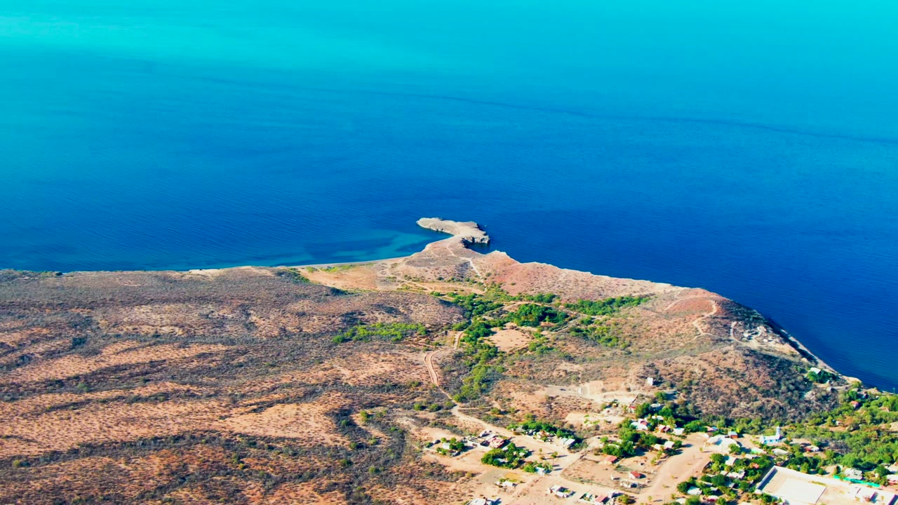 Drone view of coastline, dry terrain, roads, and clustered buildings near blue water.