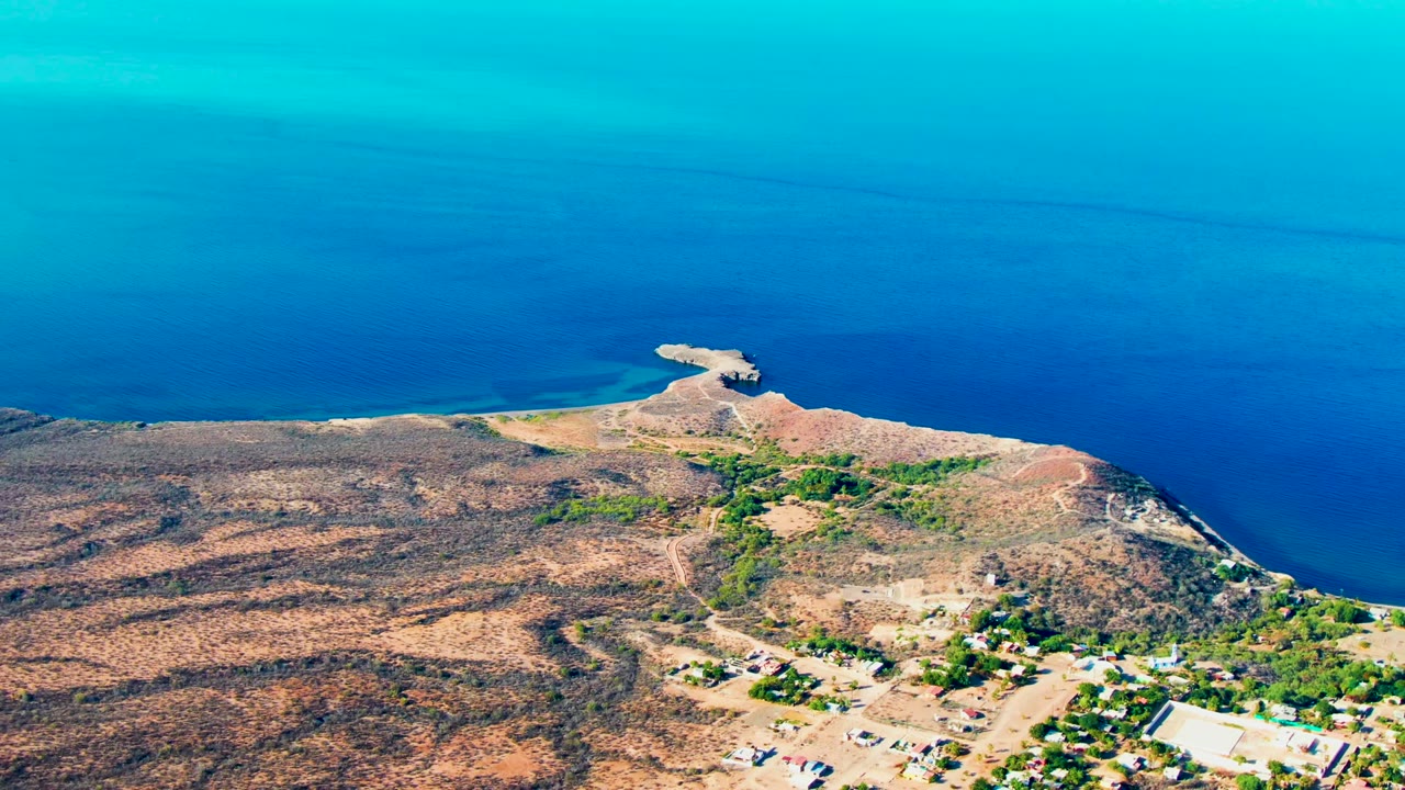 Mulege aerial frame with a broad band of blue ocean, reddish-brown shoreline, dry scrubland, road traces, settlement buildings, and no visible people.