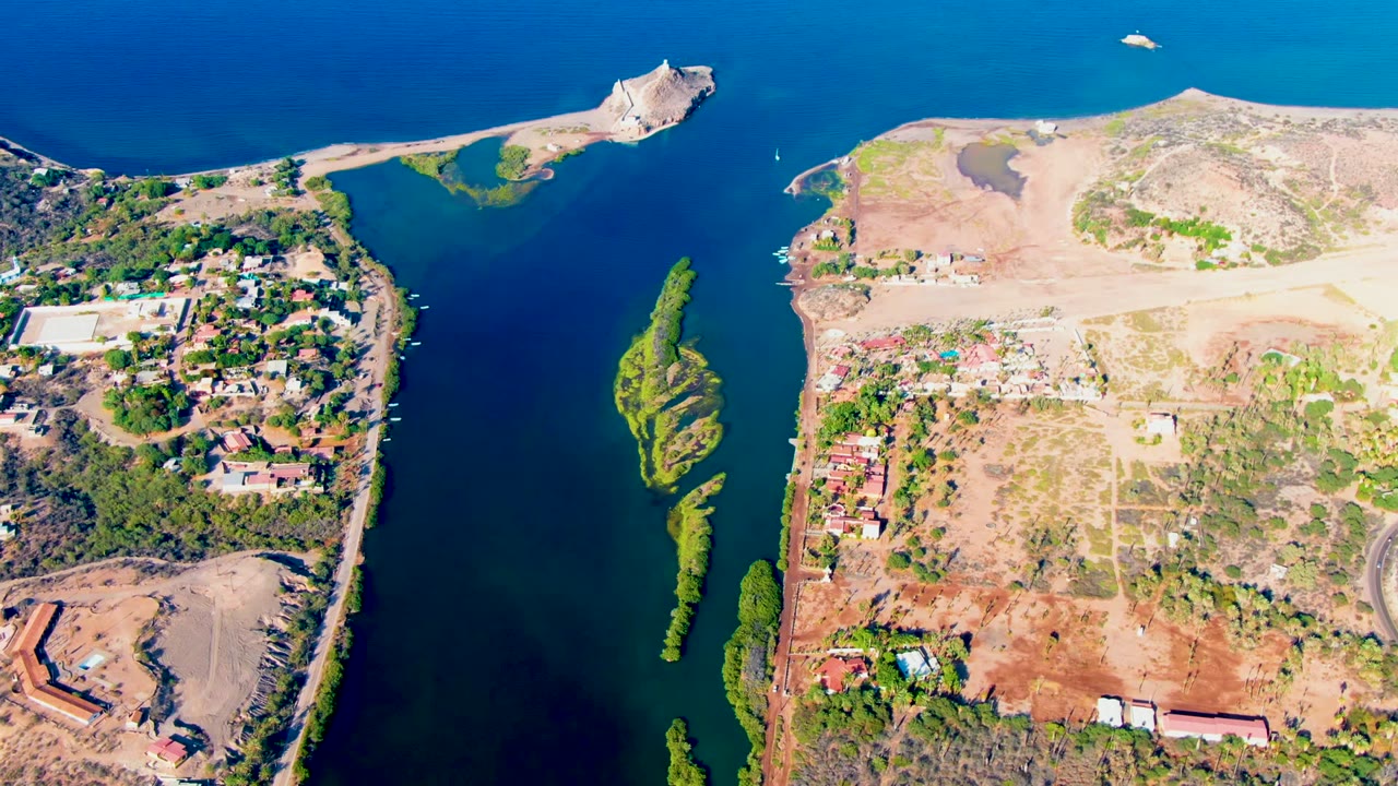 Aerial view over Mulege, Baja California Sur, showing a blue water channel, small green island-like vegetation patches, dry exposed land, scattered structures, and no visible people.