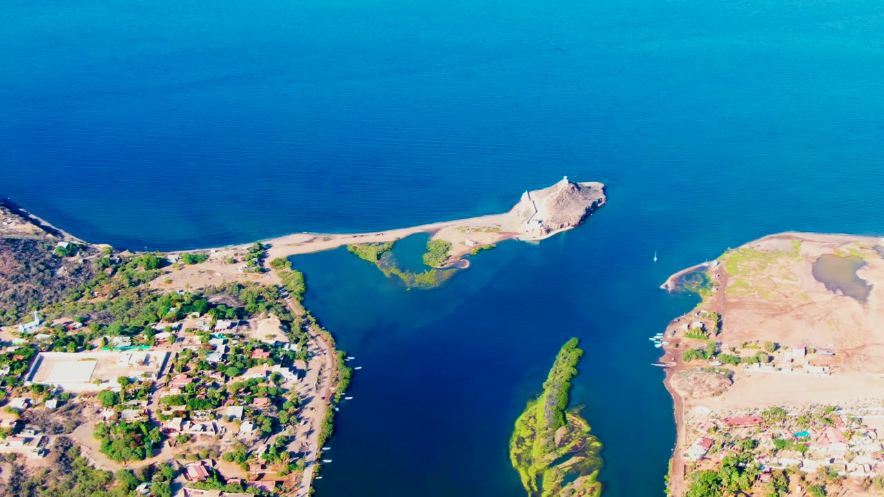 Opening aerial frame over Mulege, Baja California Sur, with blue coastal water, a narrow channel, a small green island, shoreline buildings, dry terrain, and no visible people.