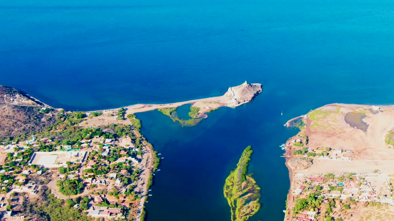 High oblique aerial over Mulege, Baja California Sur, with the green island near the lower center, built-up shoreline on one side, open tan land on the other, blue water, and no visible people.