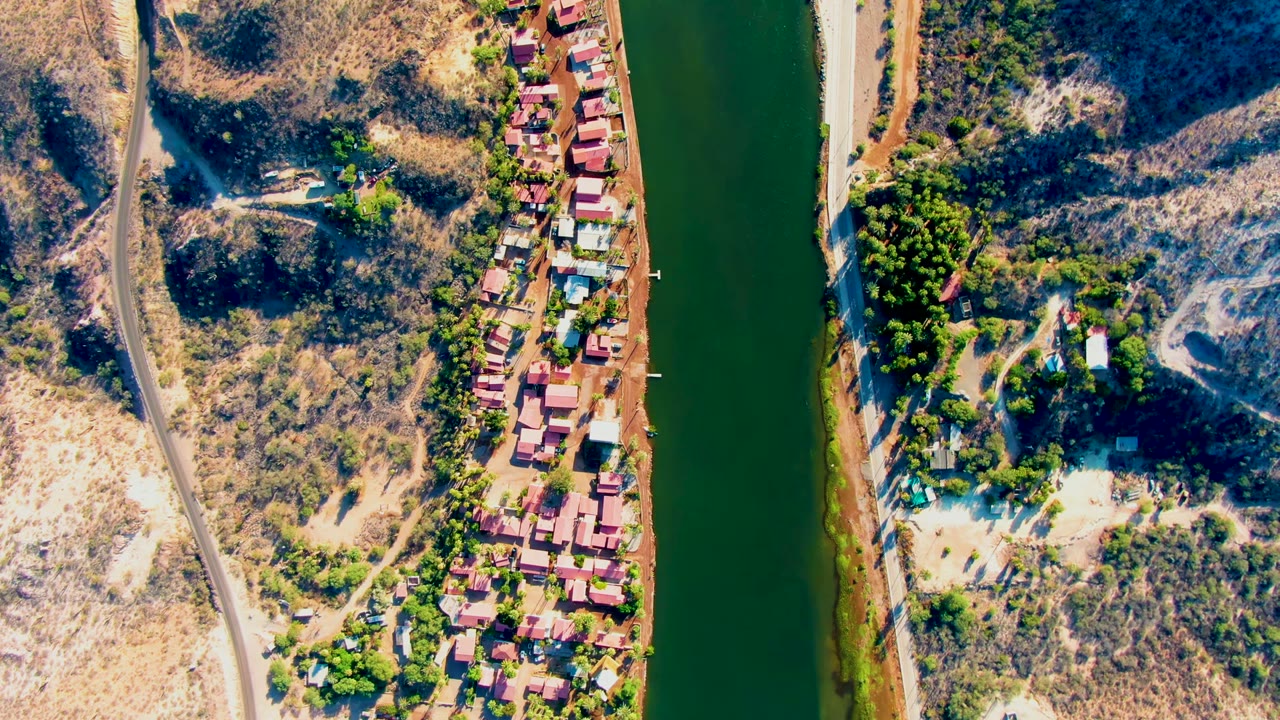 Overhead view in Mulege, Baja California Sur shows a green river, a roadside strip of small buildings, dry hillside tracks, scattered trees, and no visible people at the start of the clip.