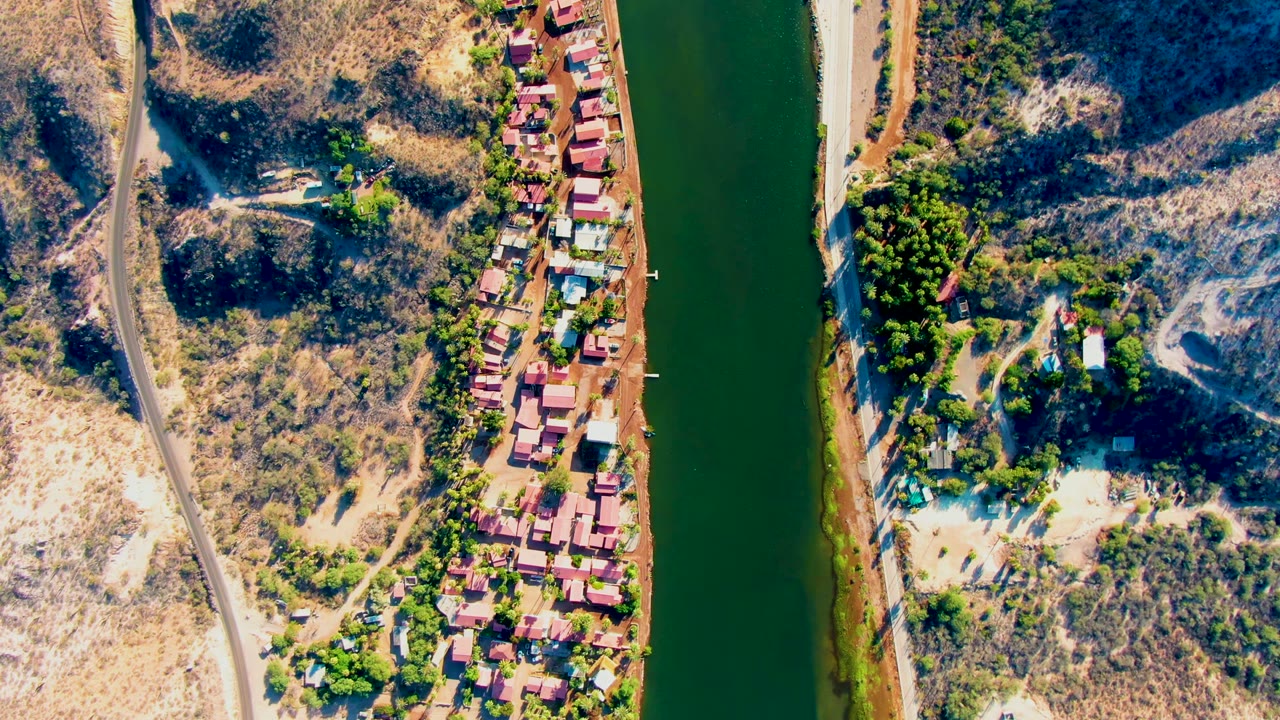 Overhead Mulege view shows the river running through frame, a road and buildings tracing the bank, pale dry ground, hillside vegetation, and no visible people.