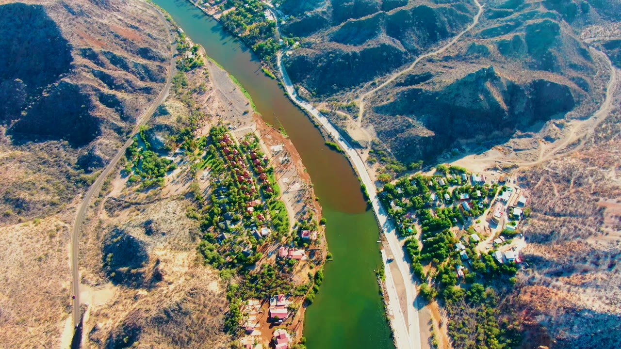 Aerial frame over Mulege, Baja California Sur, showing a green-brown river curving between dry hills, roads, trees, and residential houses with no visible people.