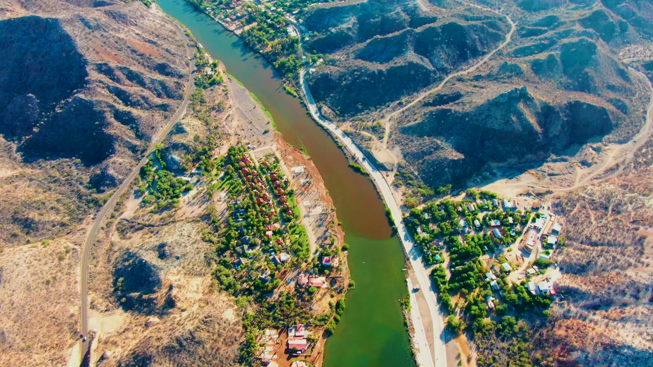 Overhead view of riverbank homes, green trees, and arid hills.