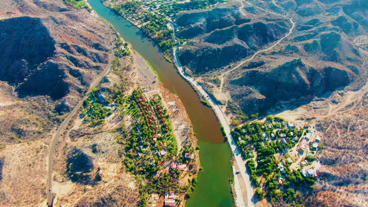 High aerial frame over Mulege, Baja California Sur, showing the river bending past a line of homes, sparse roads, and rounded dry hills, with no visible people.