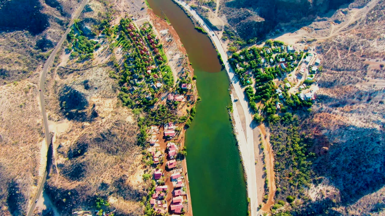 Aerial view over Mulege, Baja California Sur, showing green river water, a road with a small white car, dry hills, red-roof buildings, and no visible people.