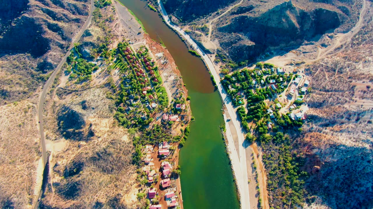 Aerial frame over Mulege with a vertical stretch of green river, roadside strip, dry ridges, trees near buildings, red roofs, and no visible people.
