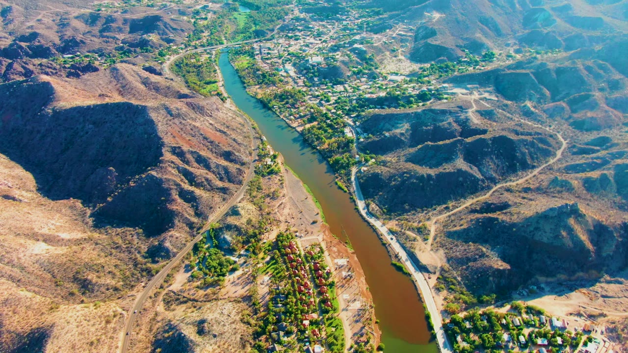 Opening aerial frame over Mulege, Baja California Sur, showing a brown-green river curving between arid hills, scattered trees, and a valley settlement with no visible people.