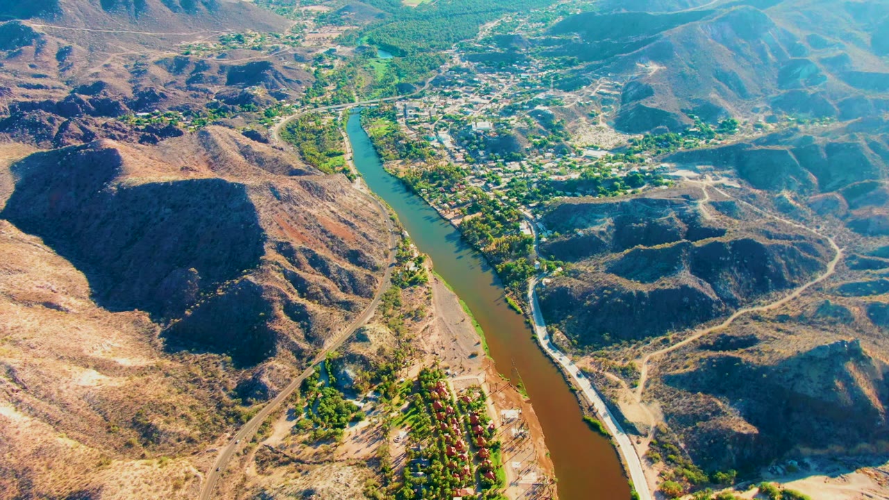 Mid-clip aerial view in Mulege, Baja California Sur, with the river curving through the center, dark hillside shadows, pale dirt roads, and no visible people.