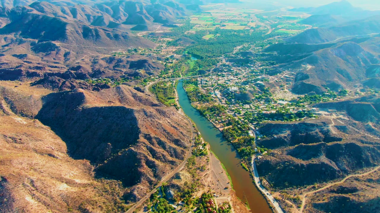 Wide aerial view over Mulege, Baja California Sur, with a green-brown river curving past a small settlement, dry mountain ridges in the foreground, distant peaks, and no visible people.