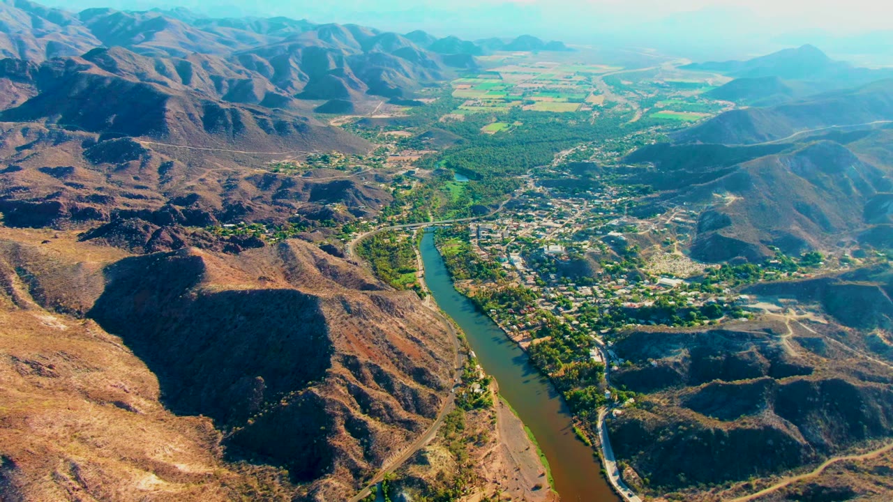 High aerial view of Mulege with the river tracing the valley edge, scattered buildings lining the bank, sunlit dry slopes, green patches, and no visible people.