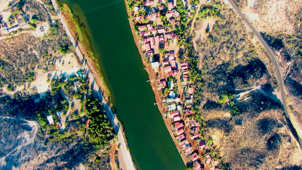 Top-down aerial frame in Mulege, Baja California Sur showing dark green river water, a narrow row of red-roofed homes, dry hillside terrain, scattered trees, and no visible people.