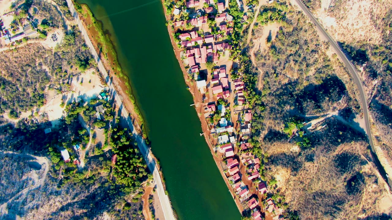 A high aerial view over Mulege shows the green river running beside red-roofed houses, pale dirt lots, scrub-covered hills, a curving road, and no visible people.