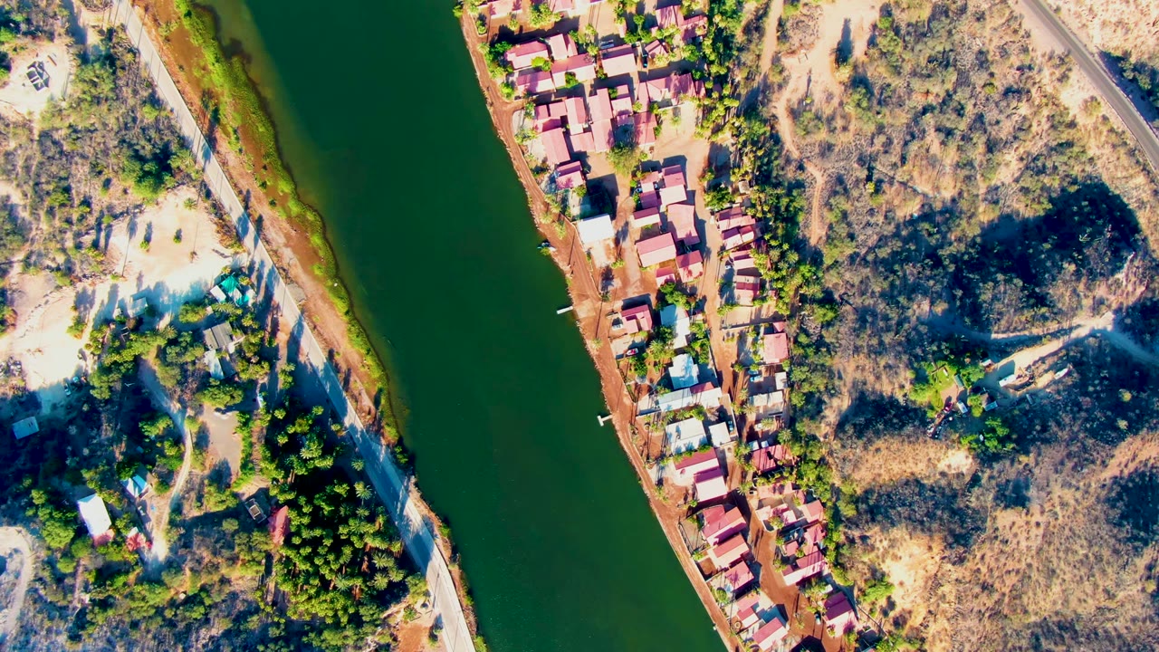 High overhead view in Mulege, Baja California Sur showing green river water, red-roofed riverside houses, a road, dry scrubland, trees, and no visible people at the start of the clip.