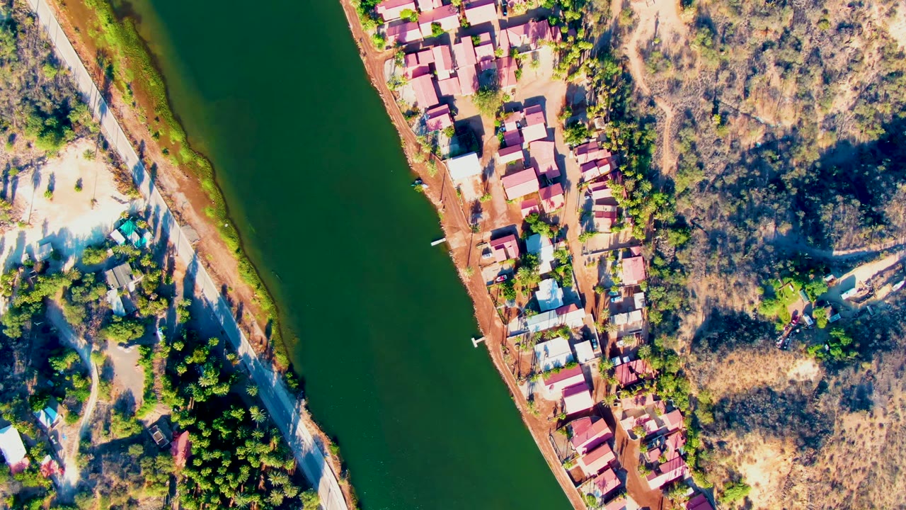 High-angle Mulege aerial view showing dark green water, a reddish shoreline, closely spaced houses, dry brush, scattered trees, and no visible people as the view advances along the river.
