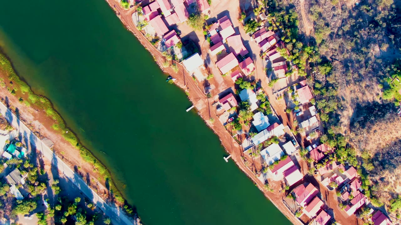 High aerial view in Mulege, Baja California Sur showing green river water, red-roofed houses along a dirt bank, dry scrubland, and no visible people.