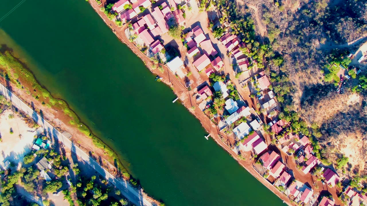 Overhead Mulege frame showing a broad green river bend, a diagonal strip of red-roofed homes, dry terrain, scattered trees, and no visible people.
