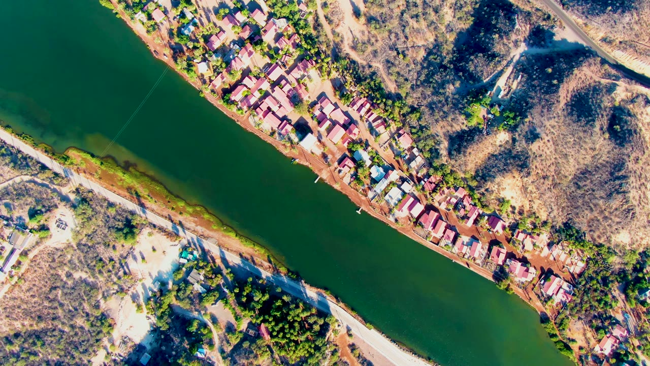 Overhead view in Mulege, Baja California Sur, showing green river water, riverbank houses, dry hills, a pale road, scattered trees, and no visible people at the opening moment.