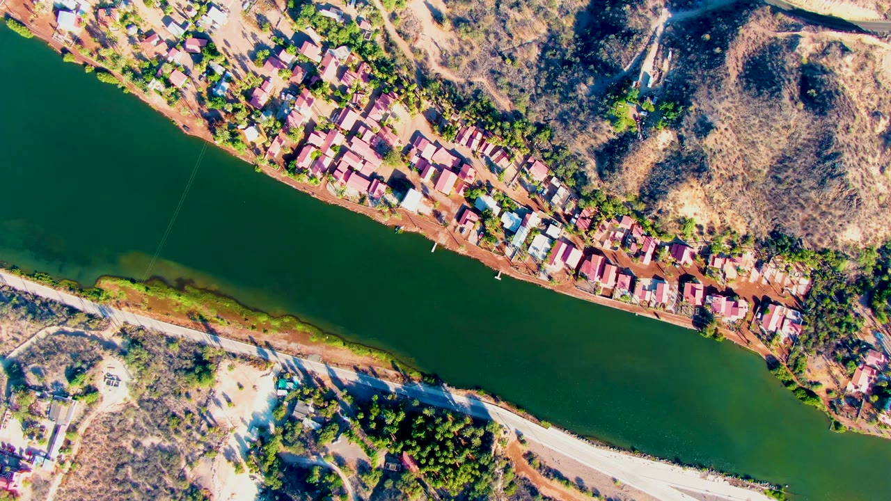 Overhead aerial frame in Mulege, Baja California Sur, showing a green river beside clustered roofs, dry hillside, sparse vegetation, and no visible people.