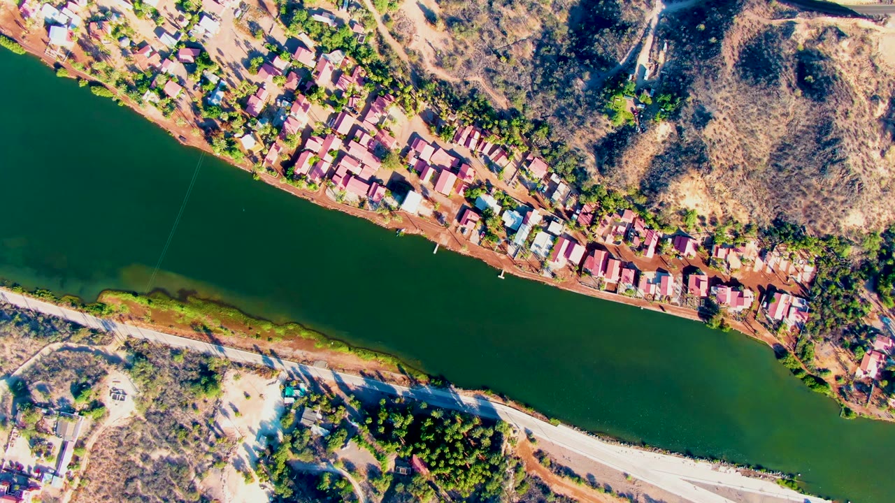 Aerial view of riverside homes, green water, dry hills, and a pale road.