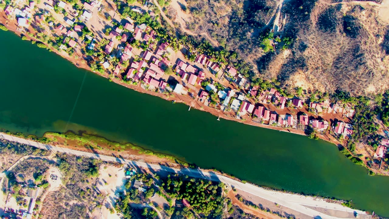 Mid-clip aerial frame in Mulege with the dark green river running below a dense line of village roofs, dry scrub-covered slopes, a pale road, and no visible people.
