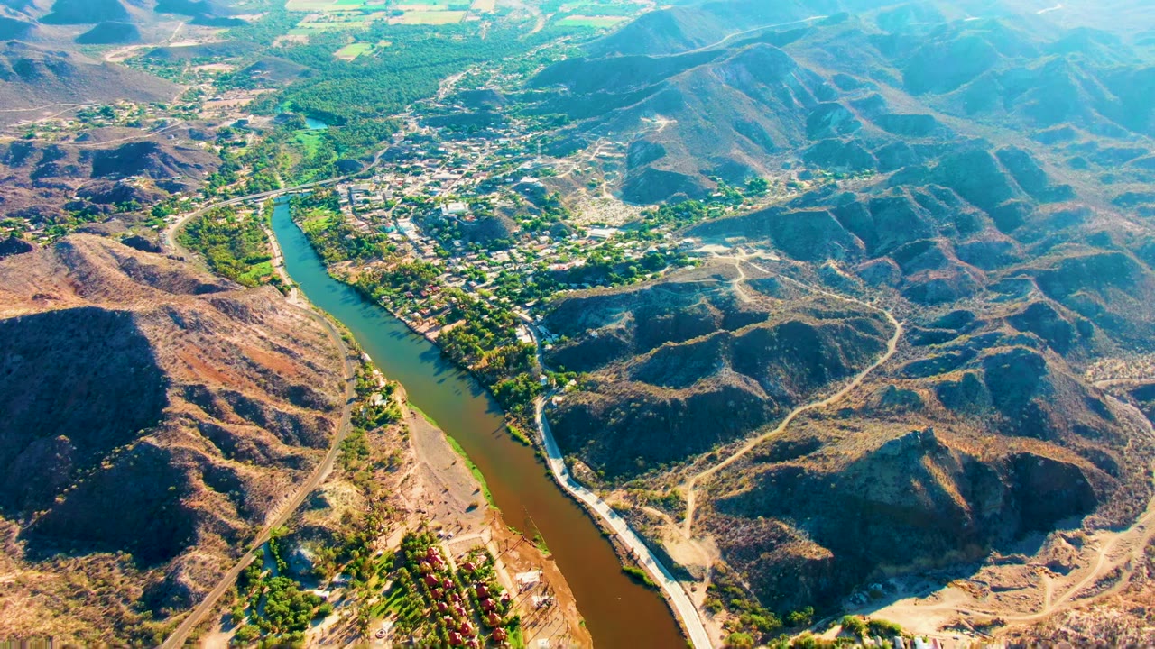 Aerial view over Mulege, Baja California Sur, with a green-brown river curving between dry hills, roads, trees, and small buildings, with no visible people.