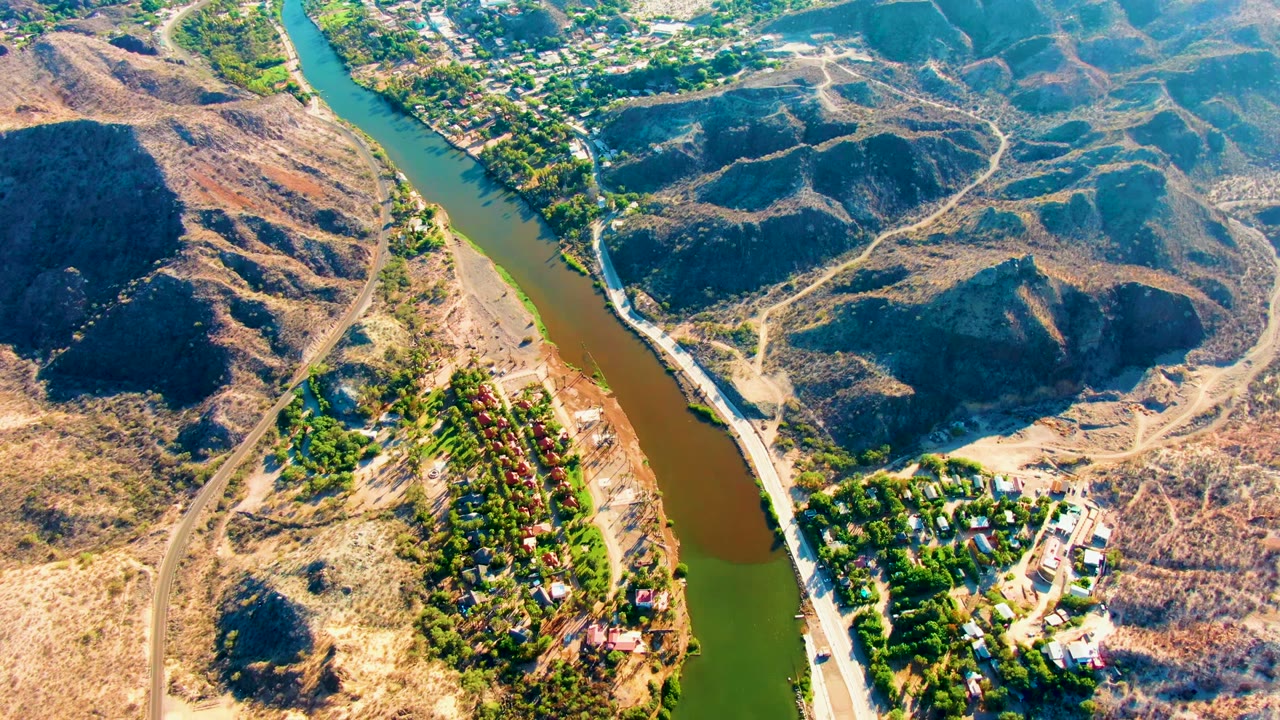 Overhead view in Mulege, Baja California Sur, with green river water, brown hills, pale dirt tracks, dense tree patches, small structures near the bank, and no visible people.