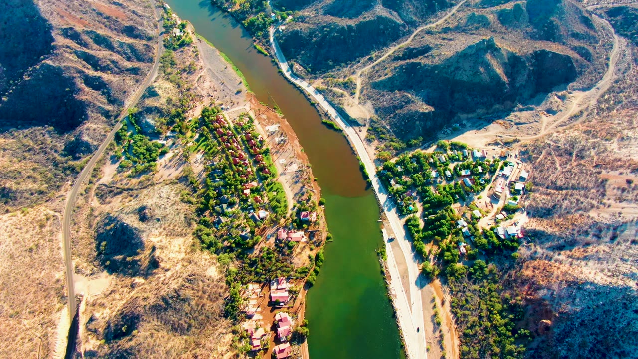 Aerial frame over Mulege, Baja California Sur, showing a pale road beside a green-brown river, dry hills, trees, scattered buildings, and no visible people.