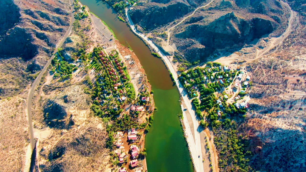 Aerial view of river, roadside vehicle, trees, buildings, and dry ridges.