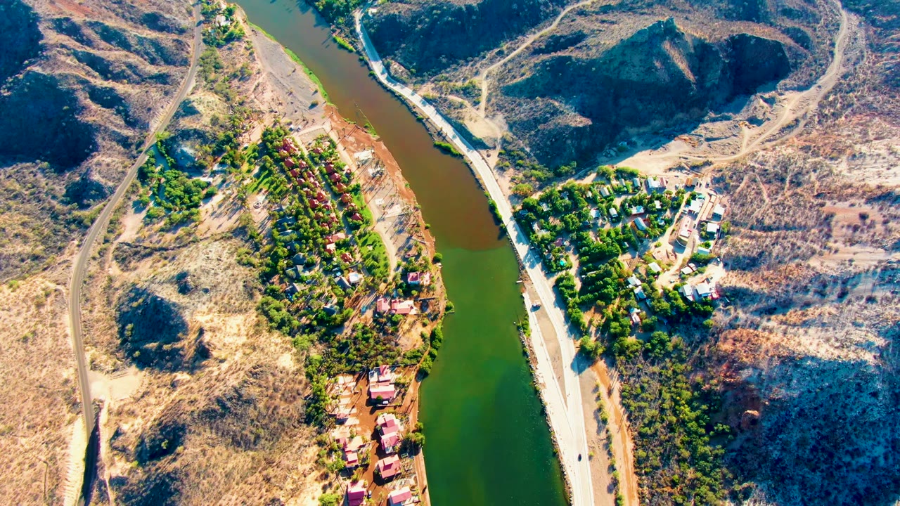 Aerial frame in Mulege showing the river edge, the parallel road, a dry hillside crossed by dirt tracks, clustered green trees, buildings, and no visible people.