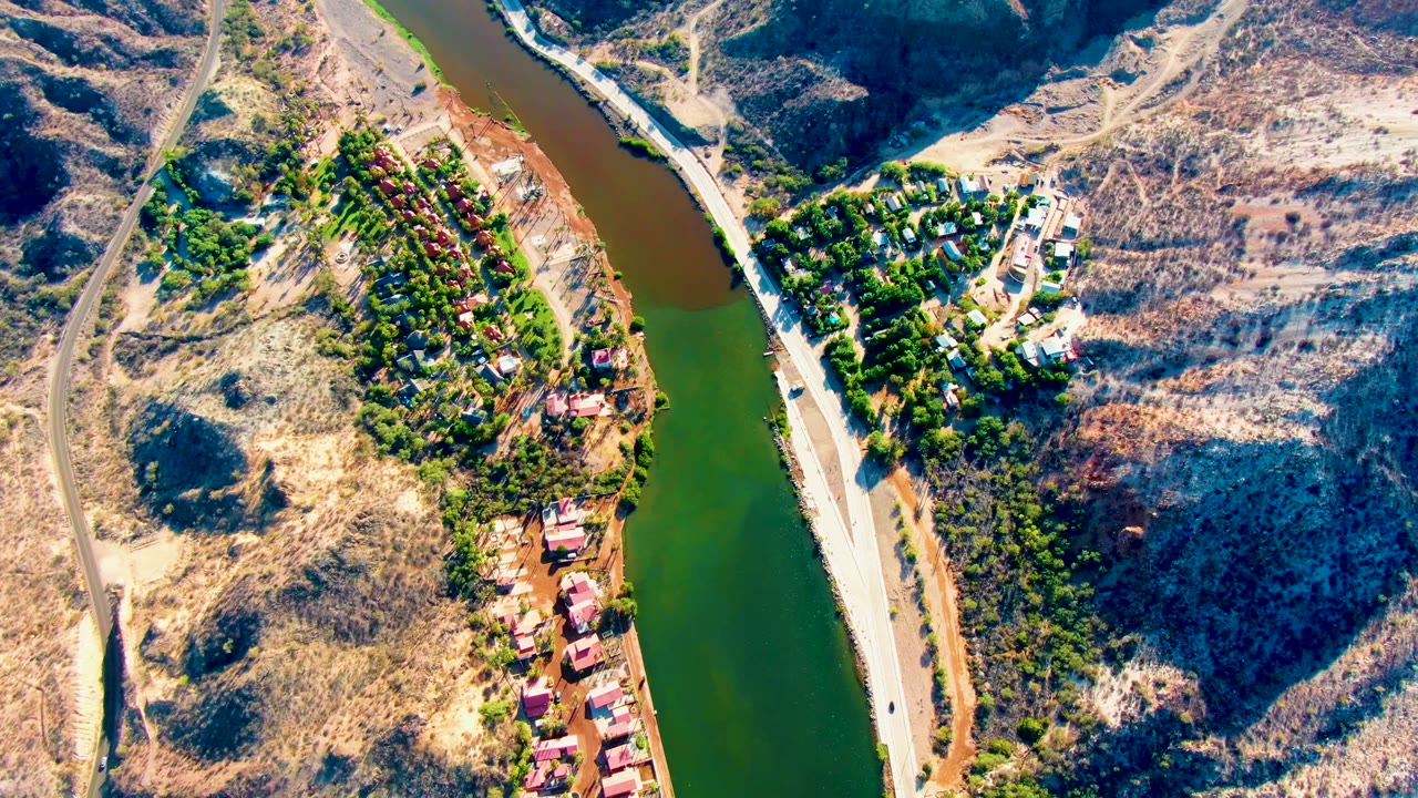 High aerial frame over Mulege, Baja California Sur, showing a green river, dry brown hills, a riverside road, clustered homes, trees, and no visible people.