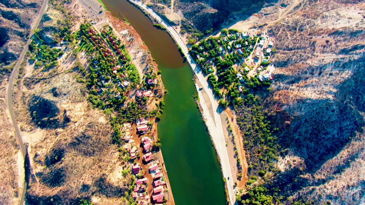 Mulege aerial frame showing the river beside a hillside settlement, a pale road along the bank, strong sun on dry slopes, deep hill shadows, and no visible people.