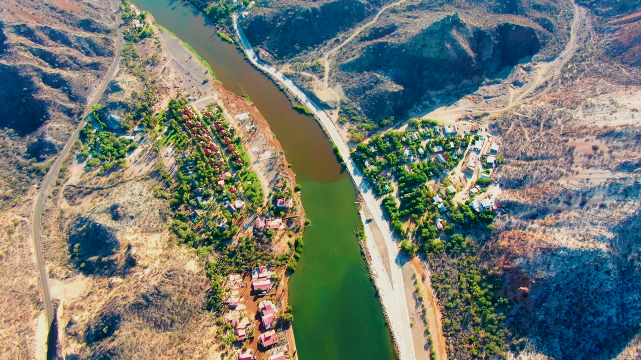 High aerial frame over Mulege, Baja California Sur showing a green-brown river bend, parallel roads, dry hills, trees, and scattered small buildings, with no visible people.