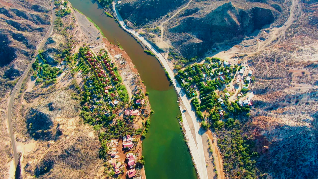 Drone-height view over Mulege with the river running vertically through the frame, dry hillside on one side, a bank road, trees, and small structures, with no visible people.