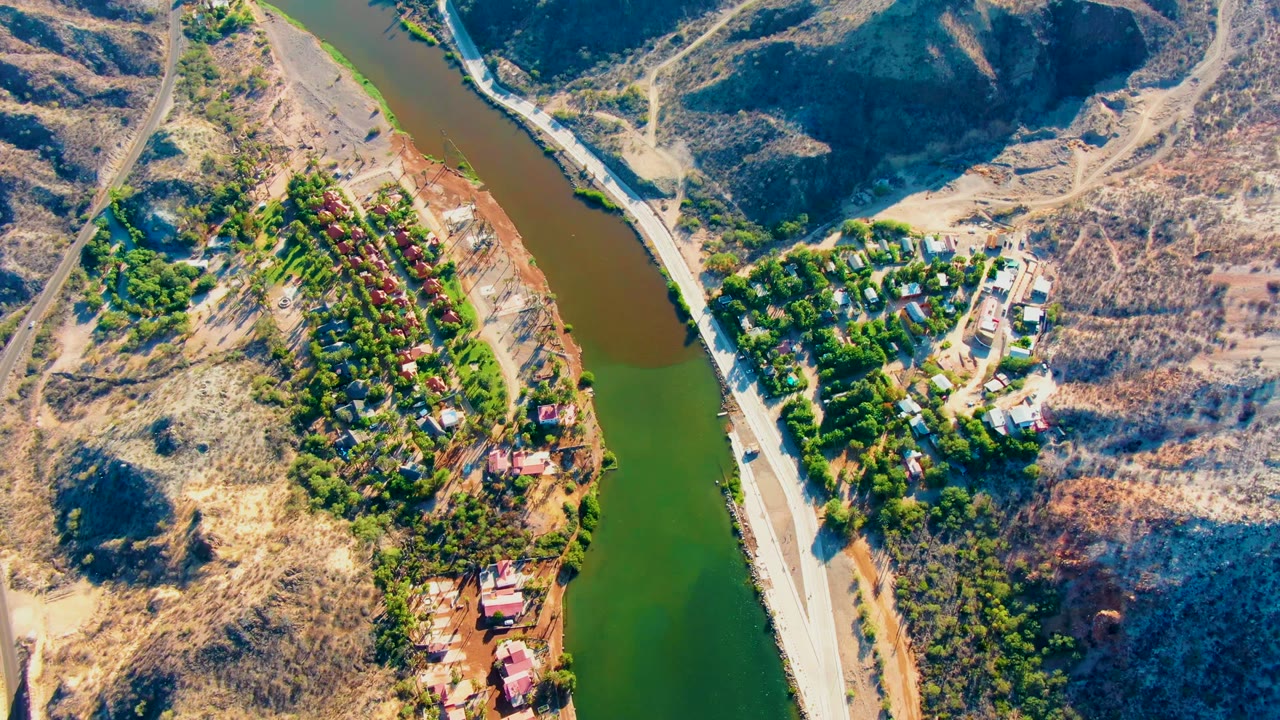 Aerial frame in Mulege, Baja California Sur with green river water beside tan arid hills, a pale road, dense trees, and small buildings near the river, with no visible people.