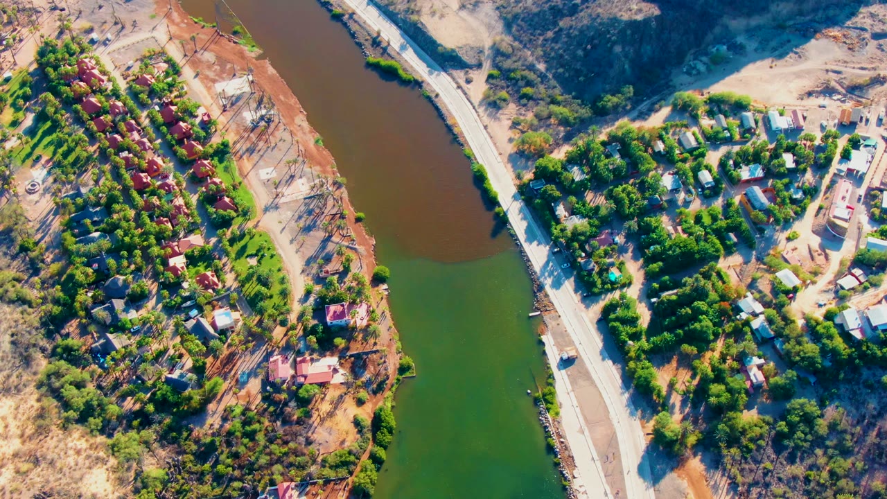 Overhead aerial frame in Mulege, Baja California Sur, showing green and brown river water beside a road, tree cover, scattered houses, dirt lots, and no visible people.