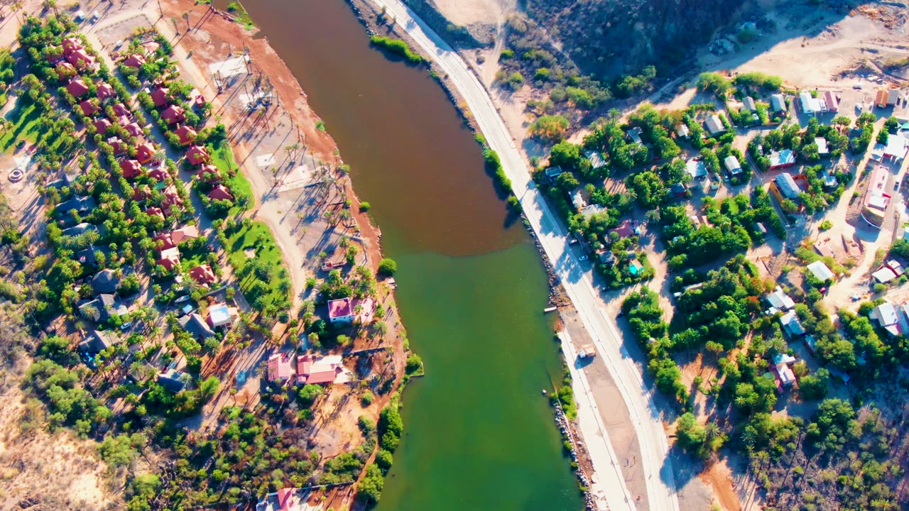 Top-down aerial view in Mulege, Baja California Sur, showing a green-brown river, a pale road, clusters of homes in trees, dry hillsides, and no visible people.