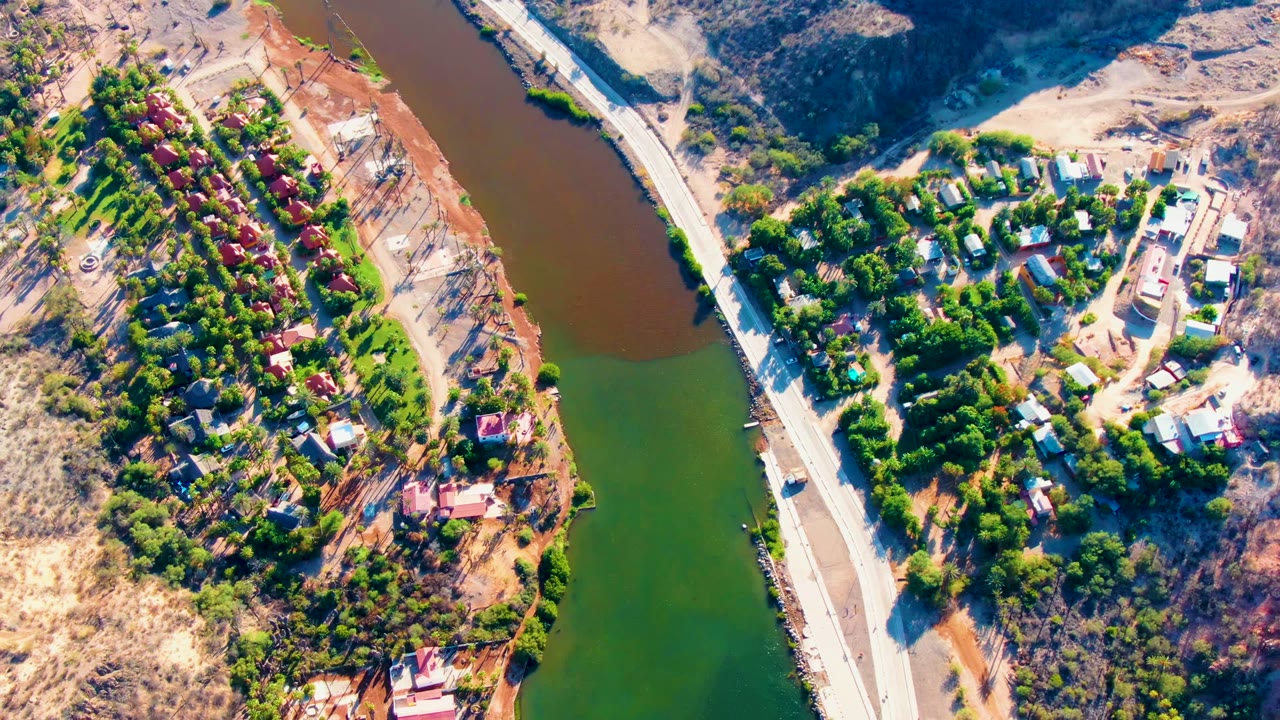 Drone-style overhead view in Mulege with red-roofed homes and trees on the left side, a road and bridge near the river, dry hills on the right, and no visible people.