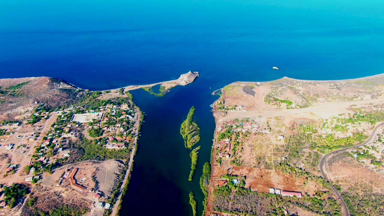Aerial frame over Mulege, Baja California Sur, showing a narrow deep blue inlet between dry land, green channel vegetation, scattered buildings, roads, and no visible people.