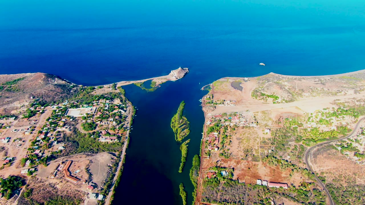 Mulege aerial frame showing the inlet mouth, a small white boat on the blue water offshore, dry hills, low buildings, roads, green vegetation patches, and no visible people.