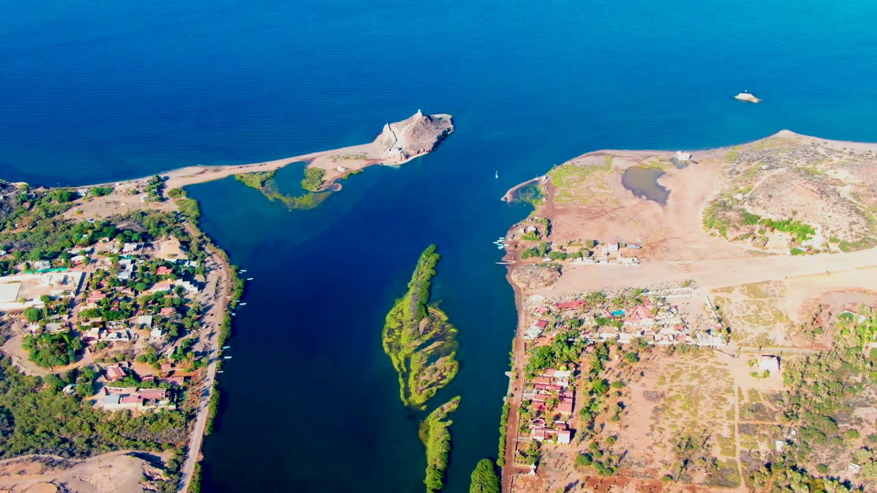 Opening aerial frame over Mulege, Baja California Sur, with deep blue water, a narrow inlet, a green island, dry exposed land, homes on both sides, and no visible people.