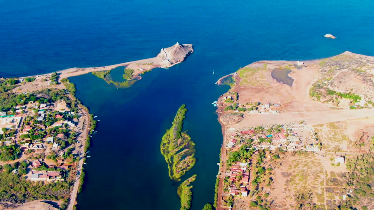 Overhead coastal inlet with exposed tan land, blue water, green island, and homes along the shore.