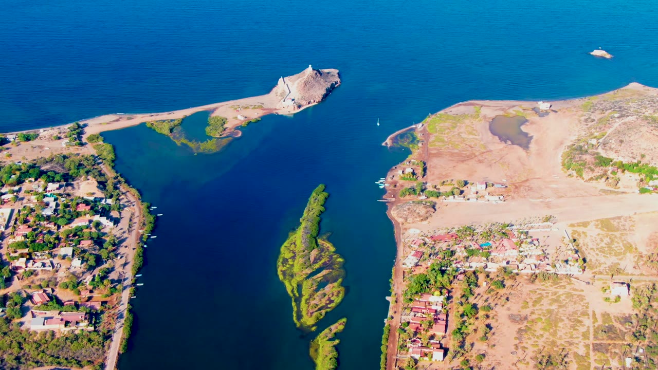Mid-clip aerial frame over Mulege, Baja California Sur, with the blue inlet running between residential banks, a narrow green island below center, exposed land on the right, and no visible people.