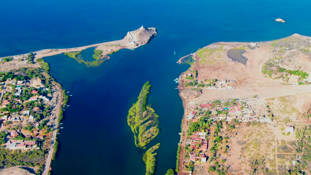 Aerial view over Mulege, Baja California Sur, Mexico, showing a blue water channel, dry shoreline, a small sailboat near the channel, a green island strip, homes, and no visible people.